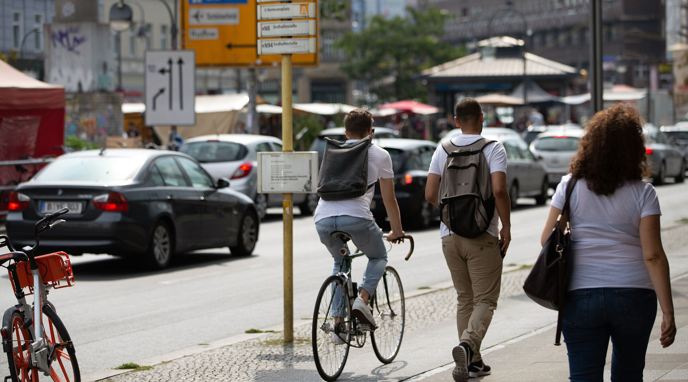 Eine Straßenszene: Ein Fahrradfahrer und zwei Fußgänger teilen sich einen gepflasterten Gehweg. Im Hintergrund parken Autos am Straßenrand.